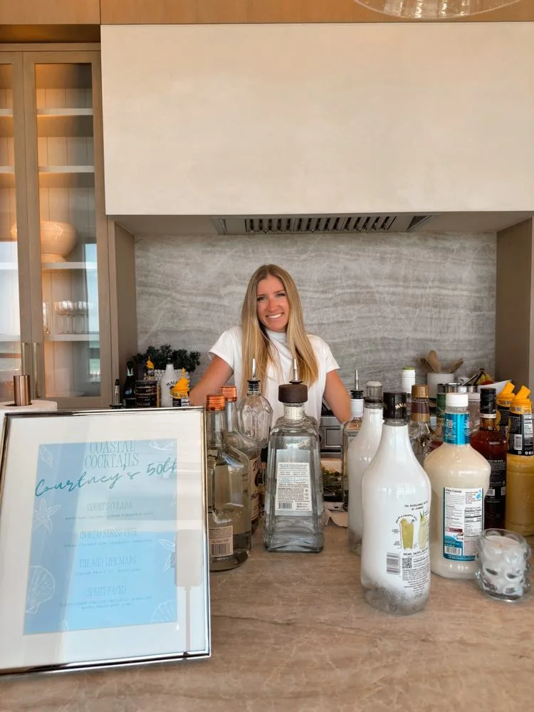 Jacque, founder of The Happy Pour, behind a fully stocked bar with a Coastal Cocktails menu