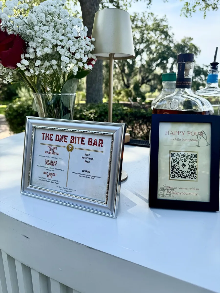 A framed custom cocktail menu next to the Happy Pour QR sign on a white bar